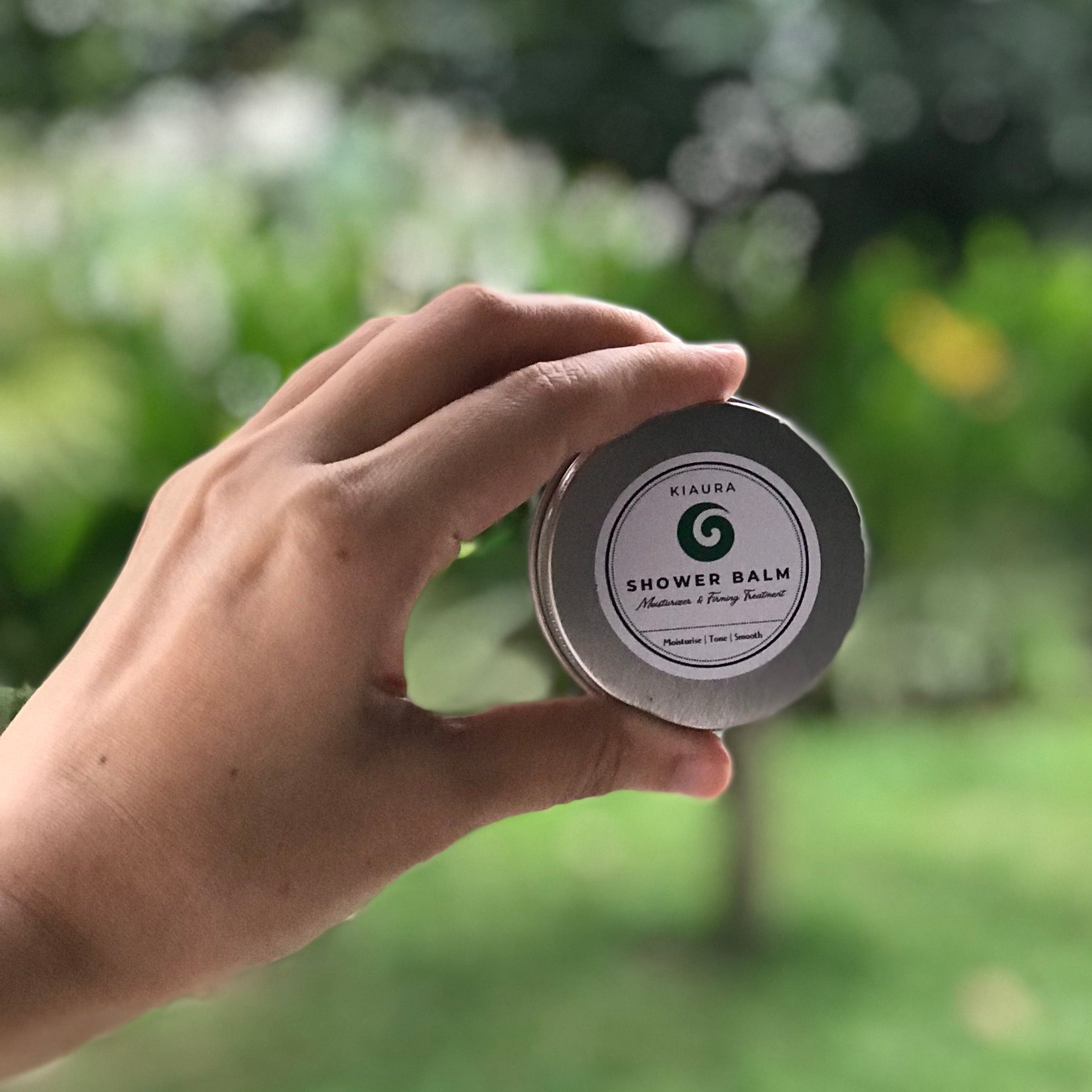 Hand holding a small container labeled 'Shower Balm' against a blurred green outdoor background